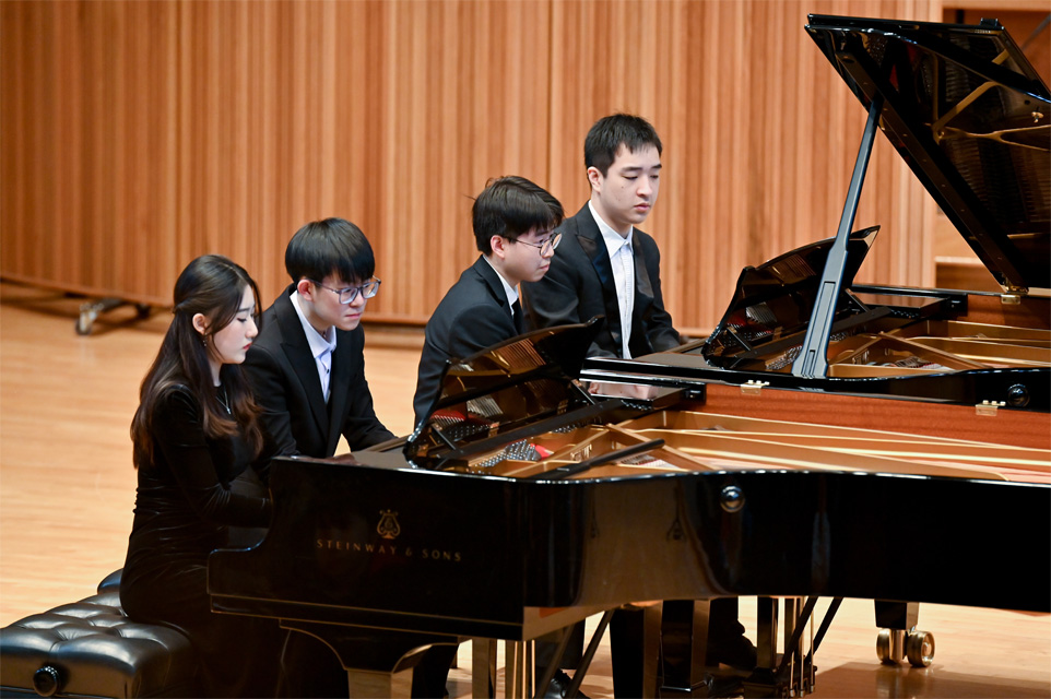 A group of students, wearing formal attire, playing in pairs on two separate pianos on stage.